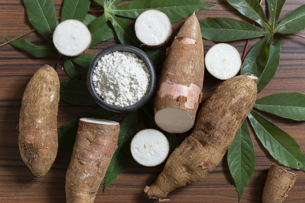 Racines de manioc entières et coupées avec bol de farine et feuilles de manioc sur table en bois